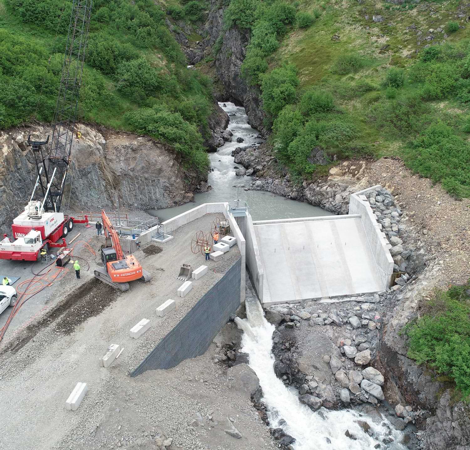 This aerial photo shows the downstream face of the diversion concrete spillway on the right, sluice channel and instream flow water release at center and pipe intake below the excavator