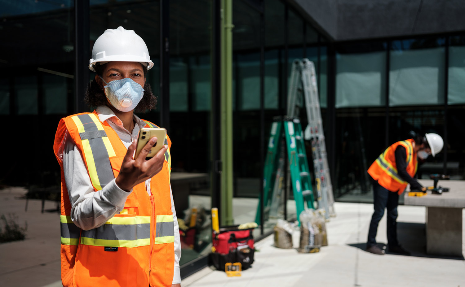 a female construction workers using a smartphone while wearing a mask, hard hat and safety vest on a site