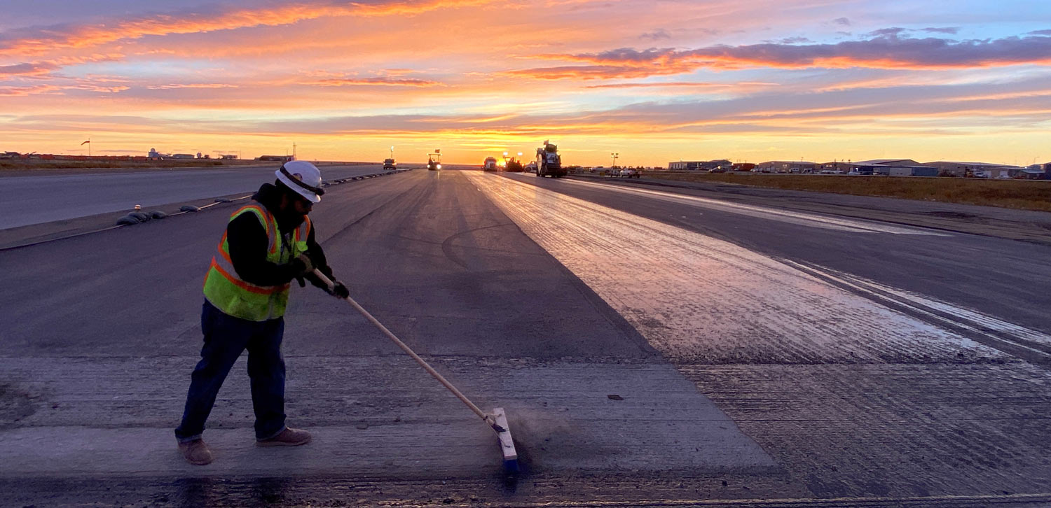 worker in a neon vest and white hard hat using a broom to get snow off a runway
