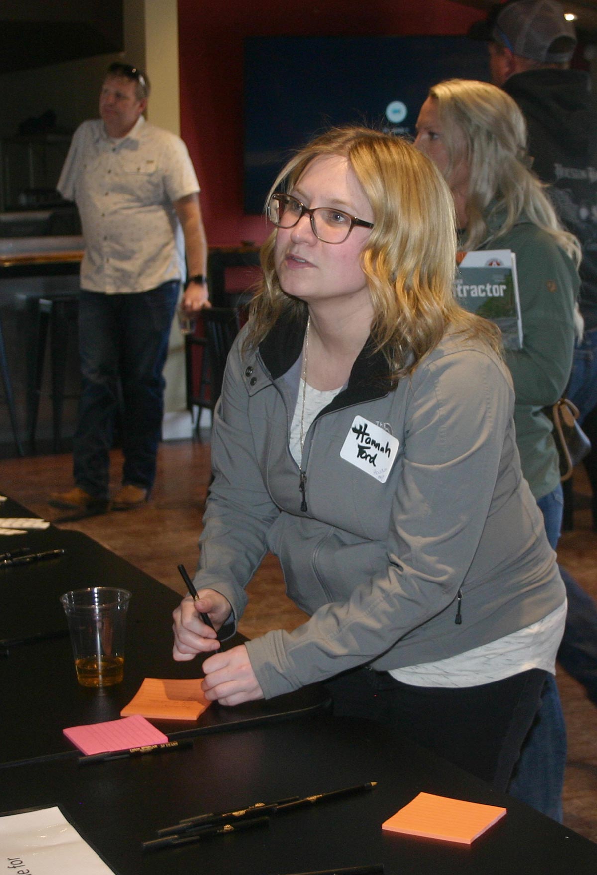 A woman examines a placard question on the wall as she has a pen in her right hand and a nearby plastic cup filled with liquid inside plus an orange sticky note ready to respond with an answer to the placard question