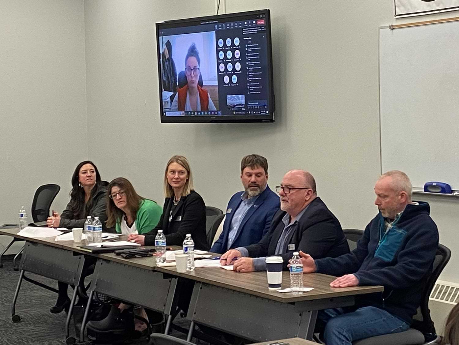 six people sit at a long table at the front of a conference room while a woman watches from a Zoom call from a television on the wall behind the long table