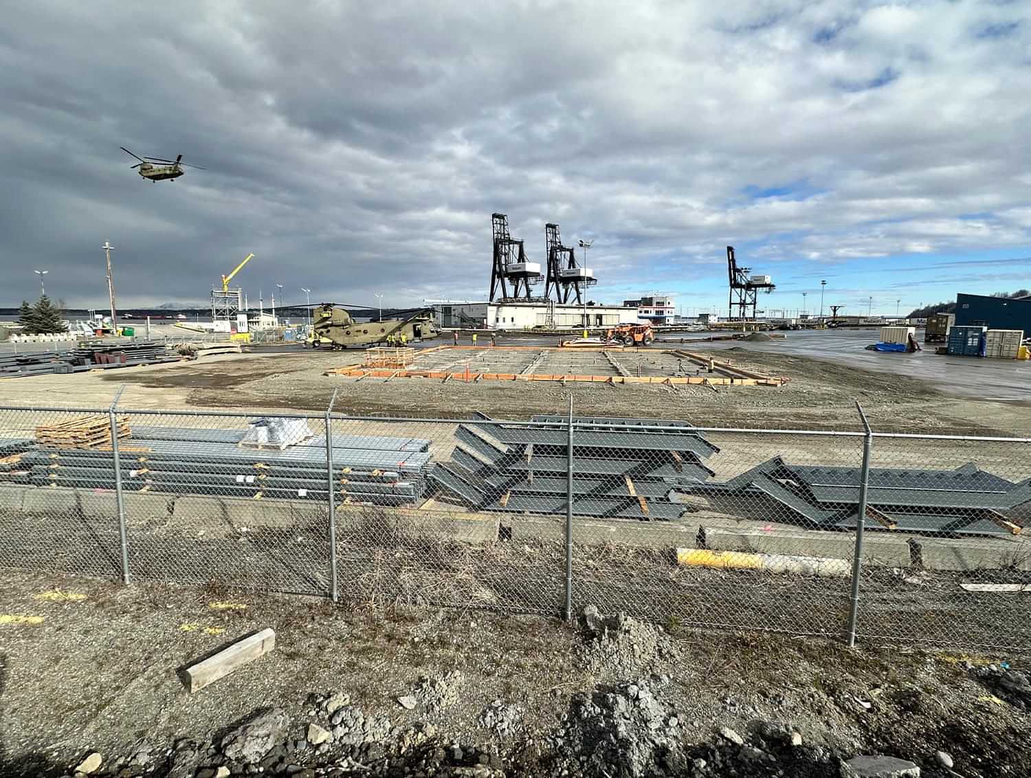 a Boeing CH-47 Chinook heavy-lift helicopter flies in the distance, over foundations and construction yard of the Port of Alaska administration building