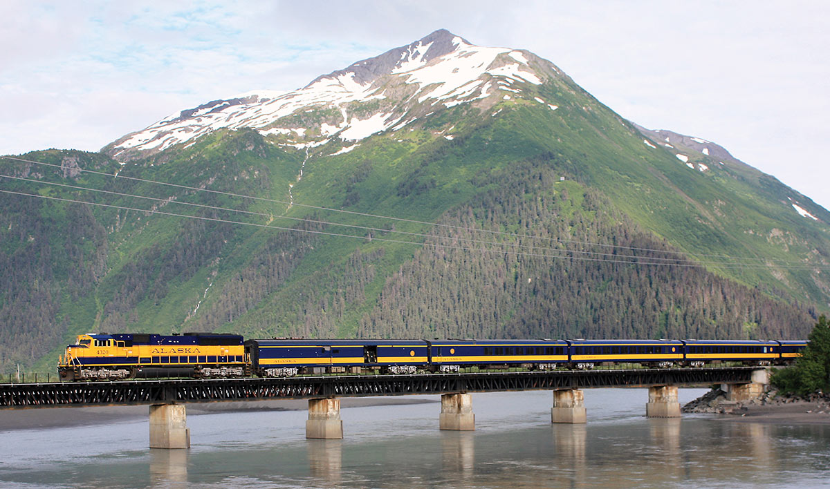 Alaska Railroad train on bridge crossing river with snowcapped mountain in background