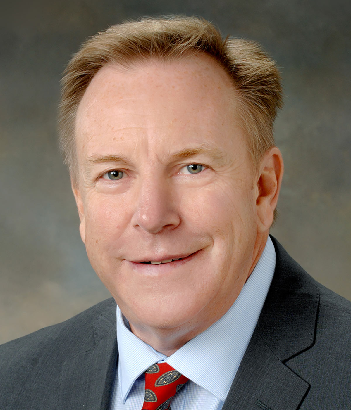 Portrait headshot photo of Tom Reilly grinning in a dark grey blazer business suit and light sky blue button-up dress shirt underneath with a red colored tie that has multi-color oval shaped pattern designs on it
