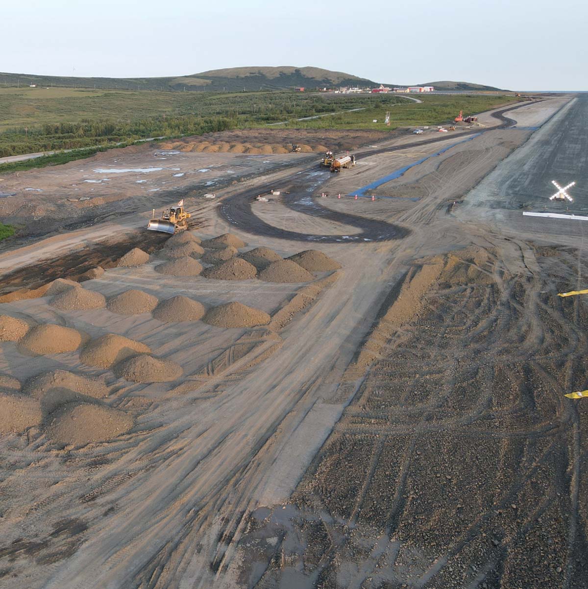 Construction site with earth movers and gravel piles near green hills.