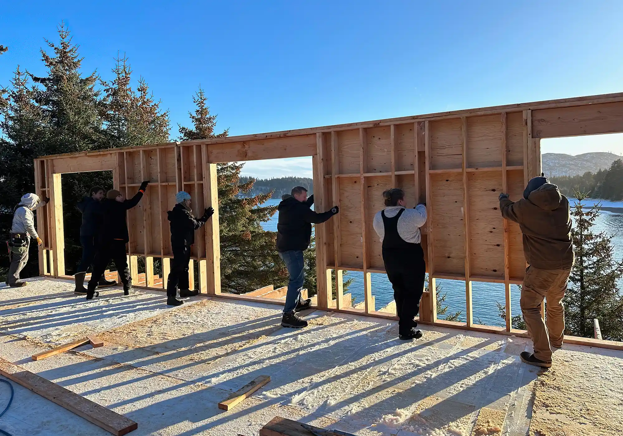 seven people work together raising a wood frame wall at a construction site near a body of water