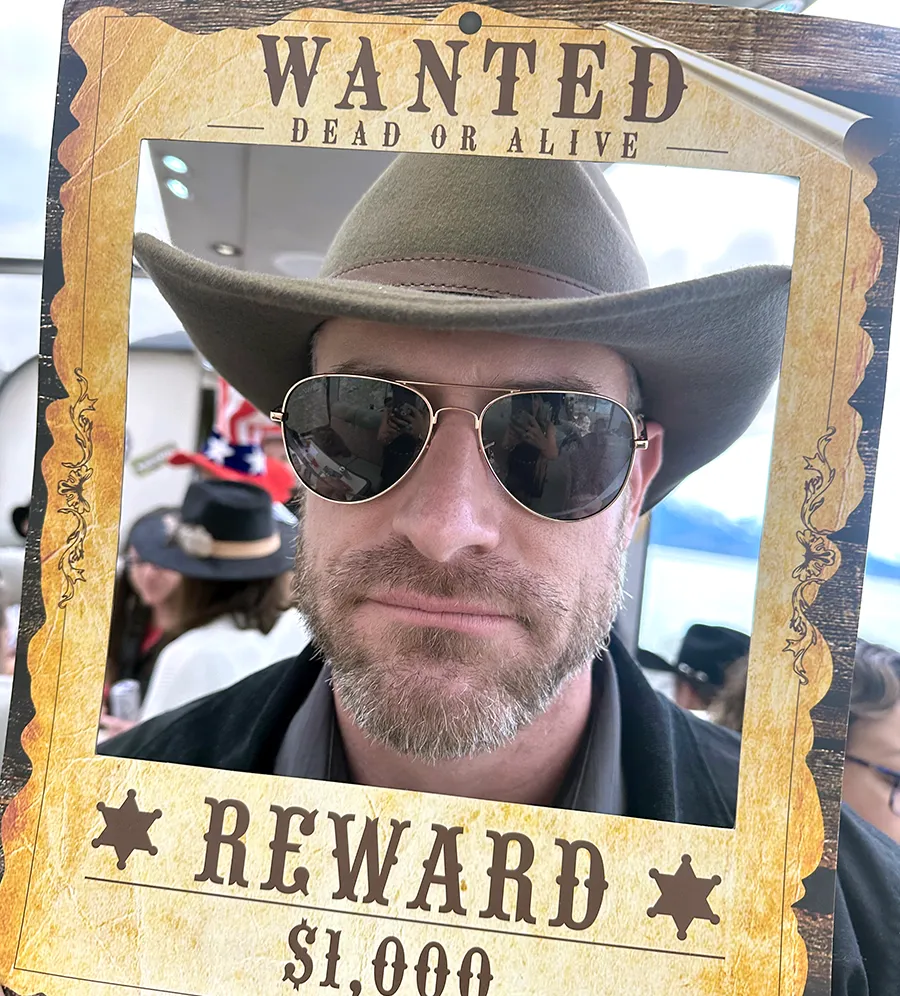 man wearing sunglasses and a cowboy hat posing with a wanted poster prop