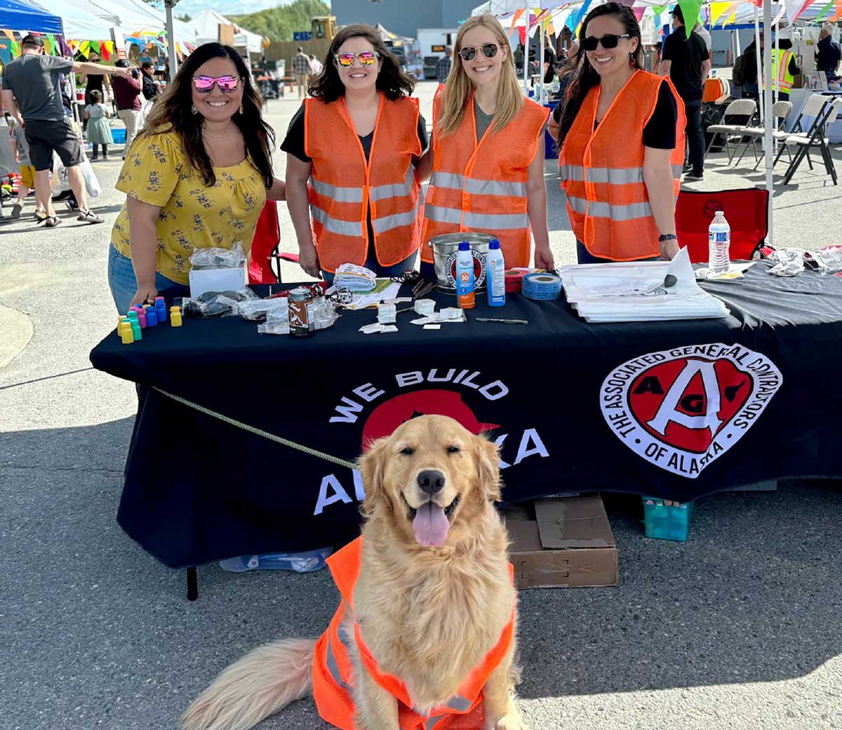 Four women and a golden retriever dog next to a black table with various items on it. The table has "WE BUILD ALASKA" and "THE ASSOCIATED GENERAL CONTRACTORS OF ALASKA" logos. Three of the women are wearing orange safety vests, and the dog is also wearing an orange safety vest.