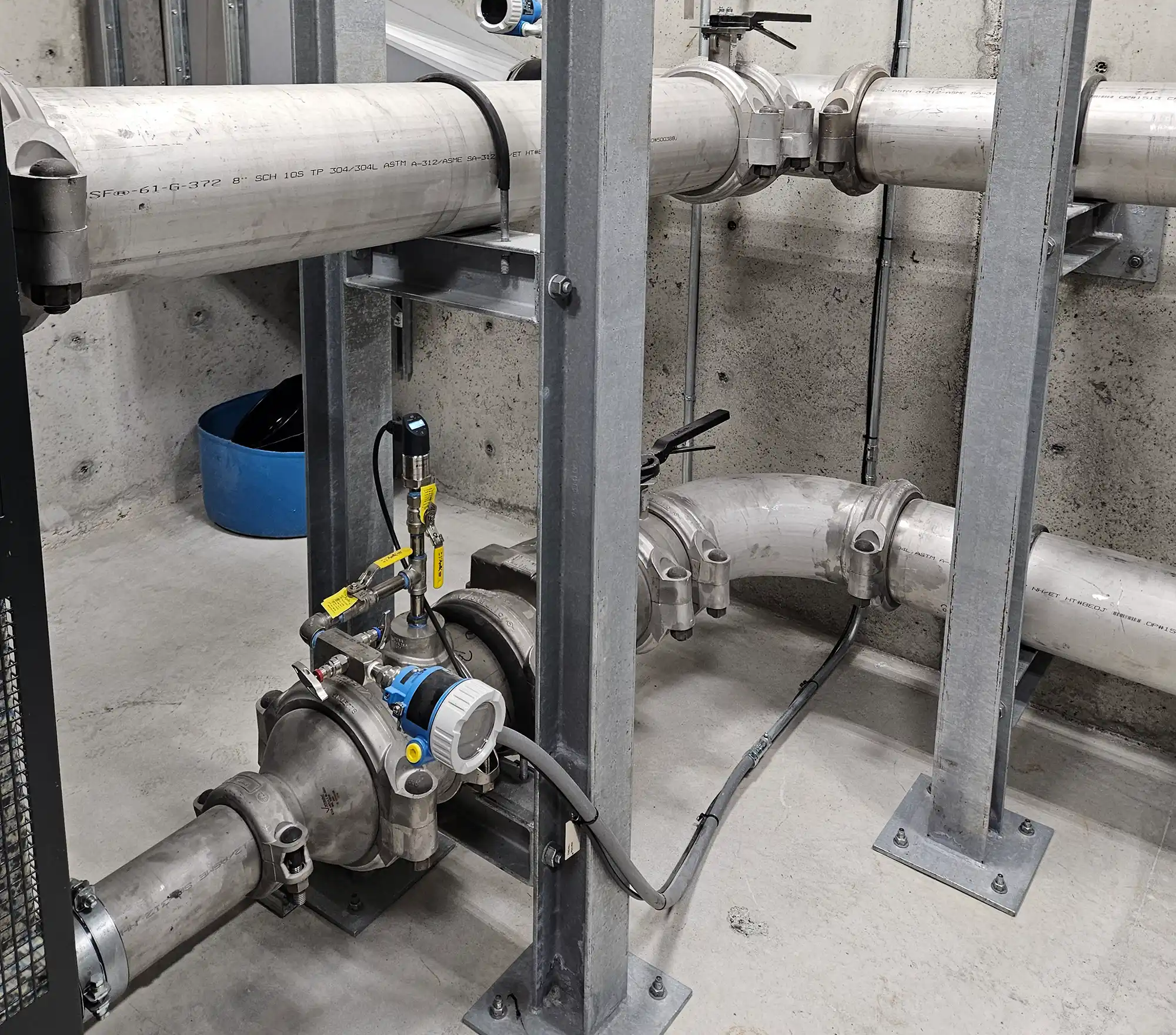 A flow meter with a digital gauge is installed on a large metal pipe, which is secured by a metal frame to a concrete floor in an industrial room.