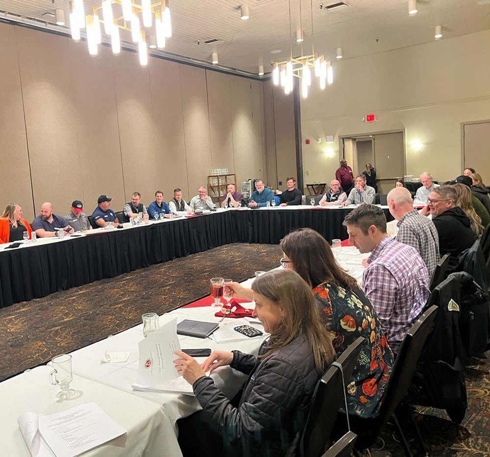 A group of people sit at long tables arranged in a U-shape for a meeting in a conference hall.