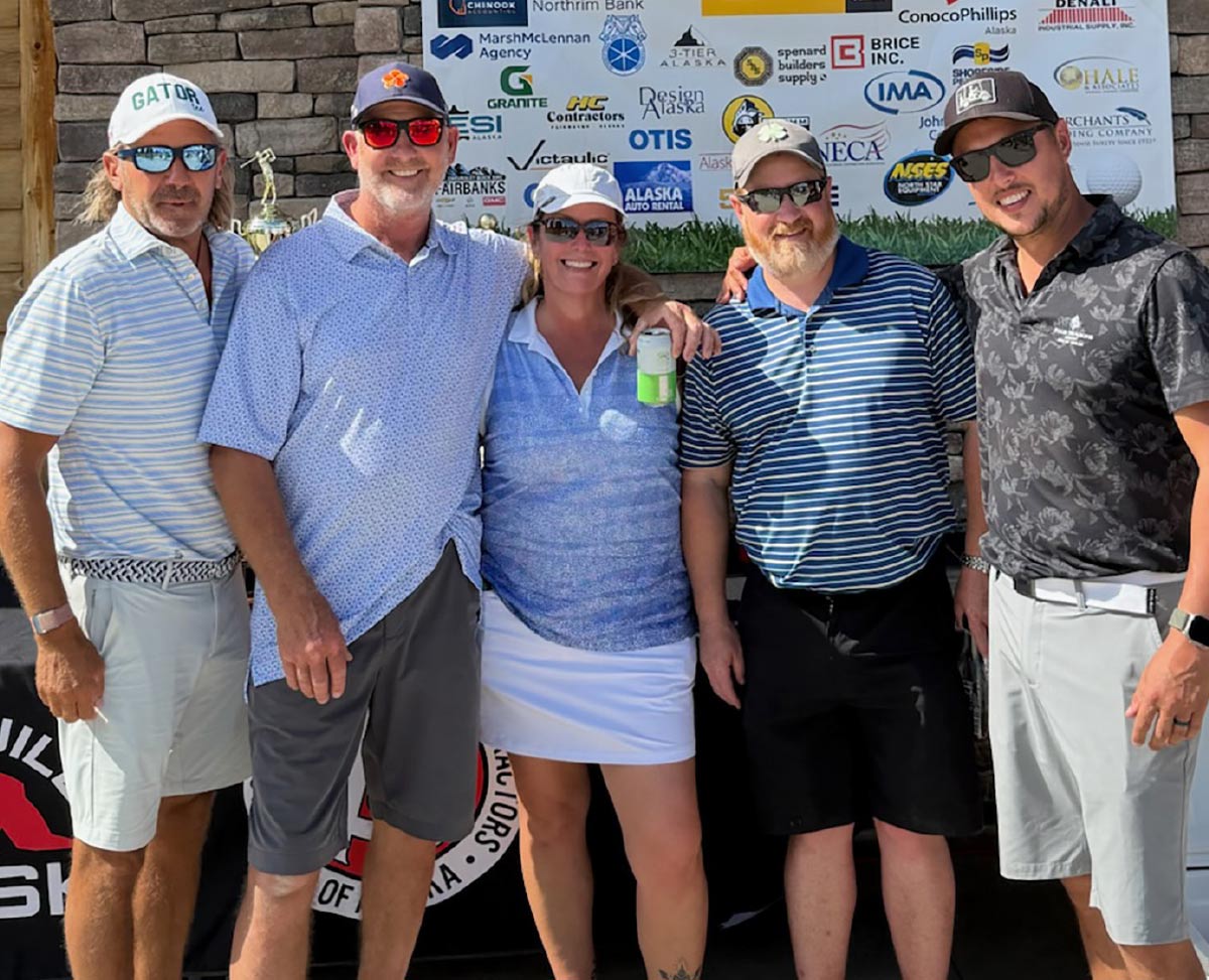 A group of five smiling golfers, one woman and four men, stand together in front of a stone wall and a "Thank You to Our Sponsors" banner.