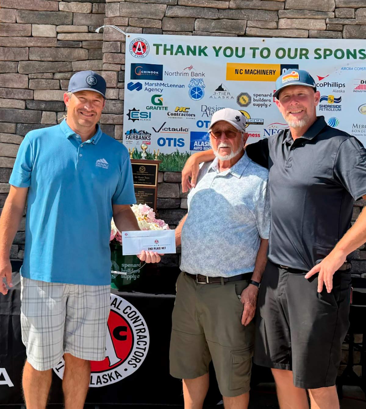 Three men, one holding a white envelope labeled "2ND PLACE NET," stand together smiling in front of a stone wall and a sponsor banner.