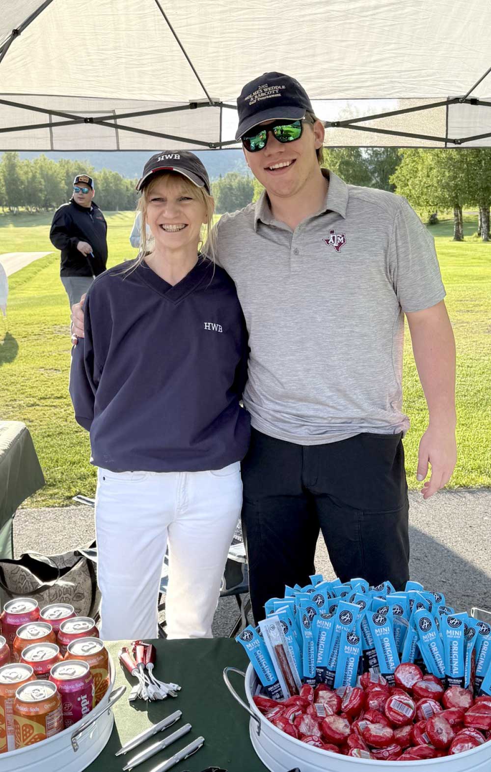 A woman and a man stand behind a table of snacks and drinks at a golf tournament, with a cooler of sodas and a bowl of mini sausages and candies in front of them.