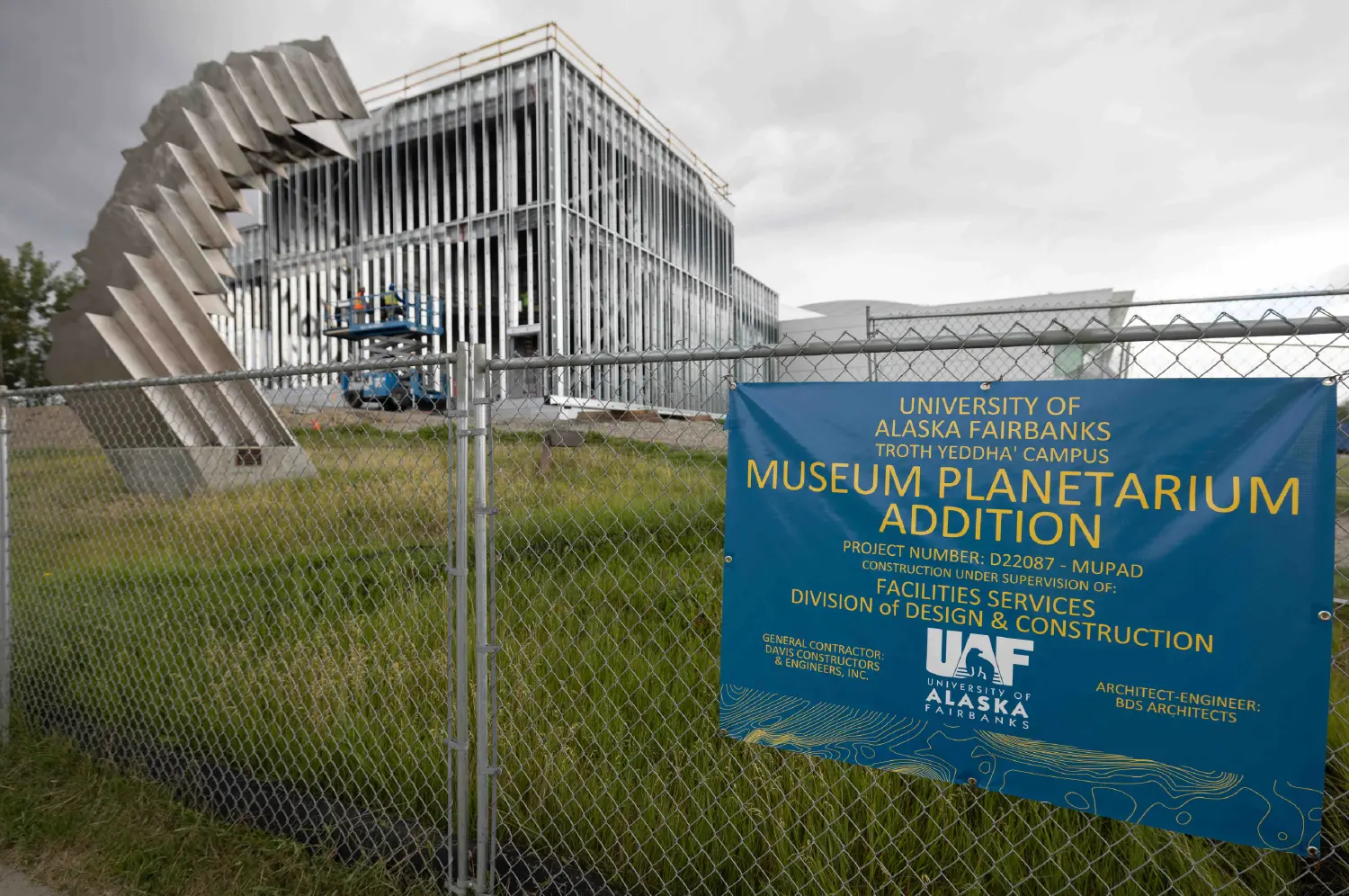 A medium-shot photo shows a blue banner with white lettering for the "University of Alaska Fairbanks Museum Planetarium Addition" hanging on a chain-link fence. In the background, a modern building is under construction. 