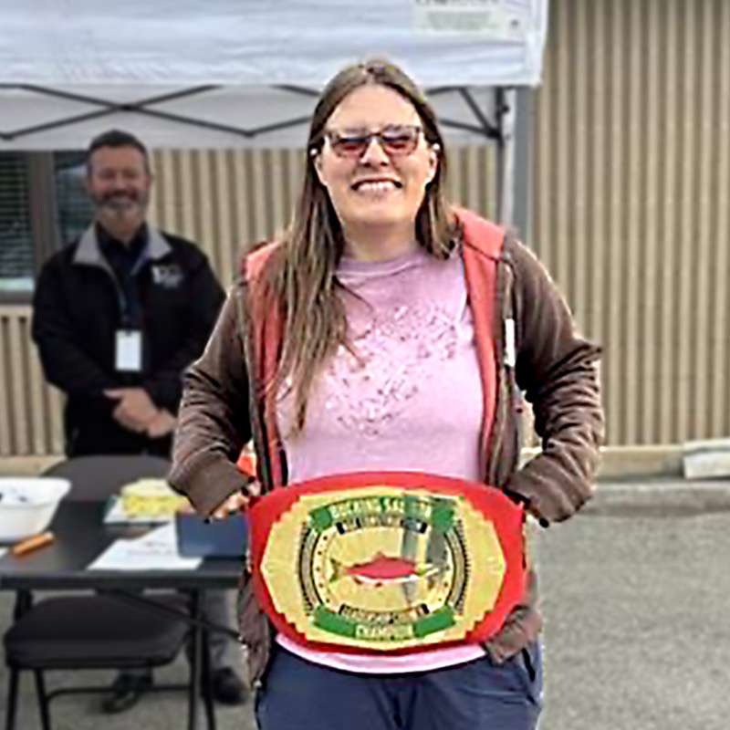 A woman smiles as she holds up a championship wrestling belt. In the background, a man with a beard stands behind a black table with a white cloth. Both are outdoors with a building in the background.