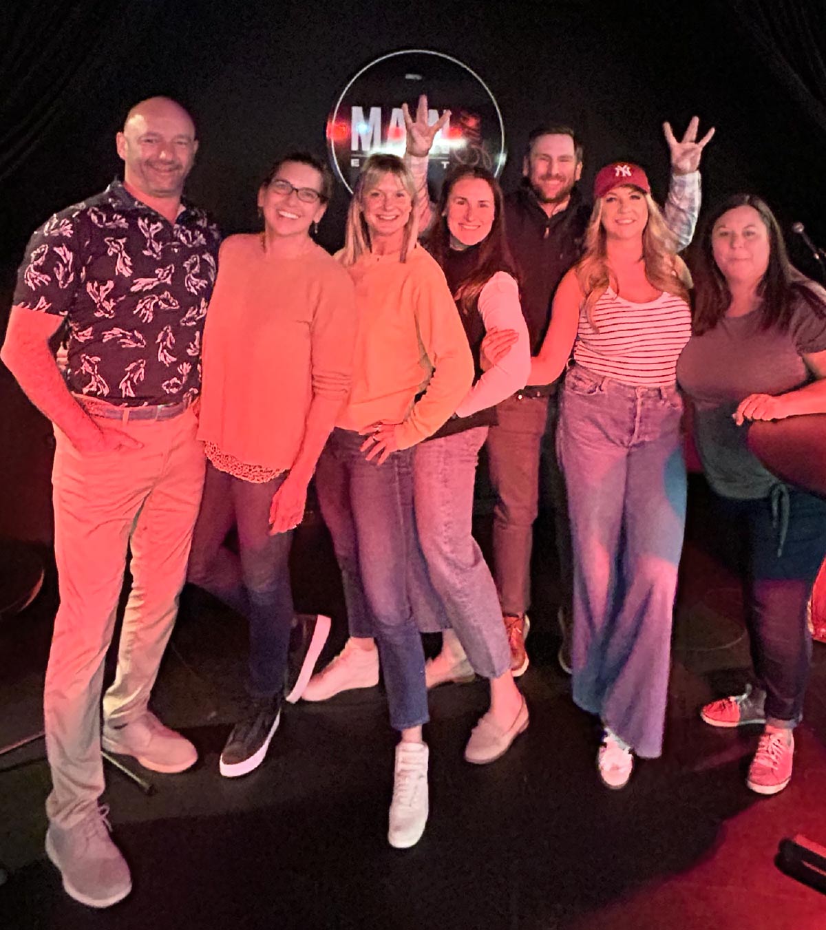 A group of seven friends stands on a stage in front of a dark backdrop with a circular logo.