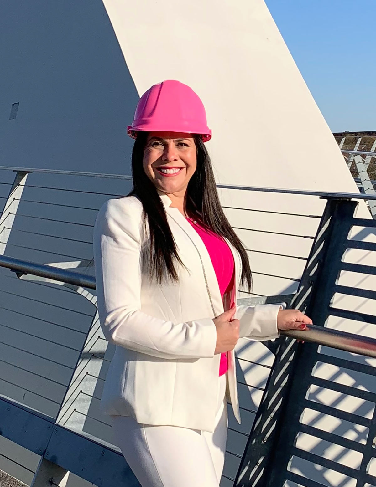 A woman wearing a white blazer, a pink top, and a pink hard hat. She is smiling and standing on a bridge-like structure with a metal railing. The background includes a modern, angular white building and a clear blue sky.