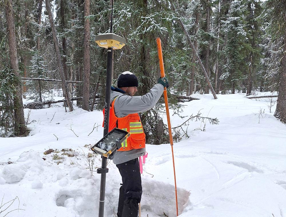 A surveyor, Brett Fillipi, wearing a winter hat, jacket, and an orange reflective vest. He is in a snowy, wooded area, holding a long orange pole with one hand while using his other hand to install a Carsonite stake into the ground. A survey monument device is mounted on a tripod nearby.