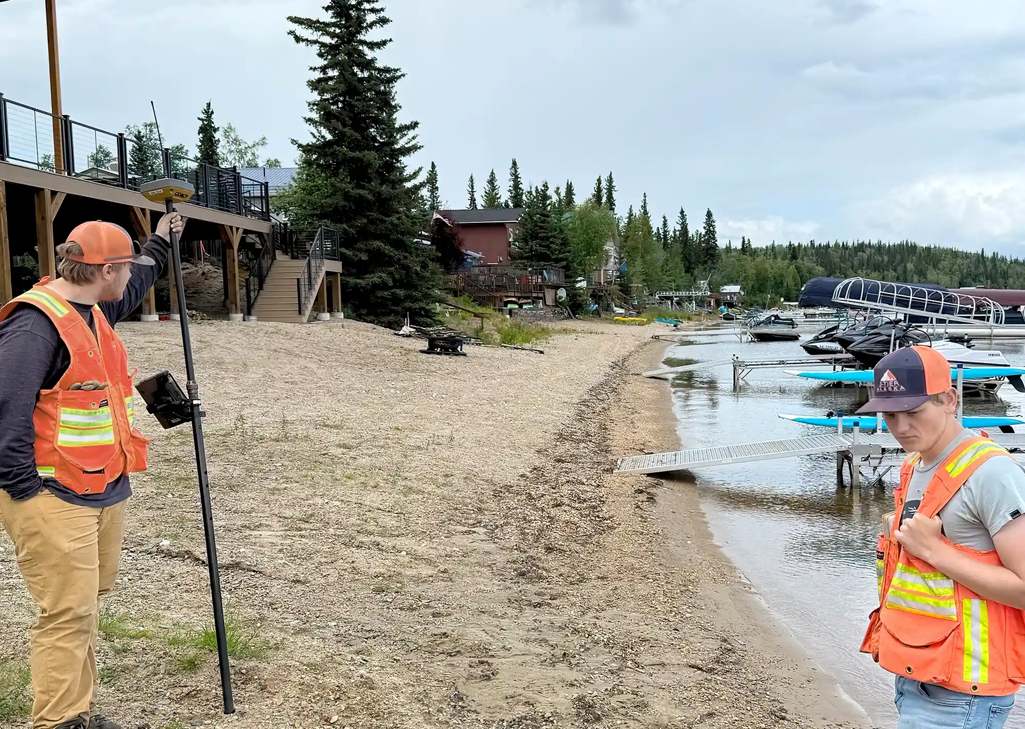 Two surveyors, Gabe Fowler and Cole Hudnall, wearing orange reflective vests and hats, standing on a gravelly beach next to a lake. One surveyor holds a long pole connected to surveying equipment, while the other looks down at the ground. In the background, there are docks with boats and a lodge building.