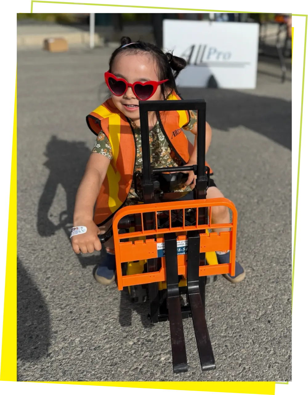 A young girl with pigtails is smiling and looking at the camera while riding a toy forklift. She is wearing a floral dress, red heart-shaped sunglasses, and a small orange safety vest. She has an event wristband on her left wrist. The scene is outdoors on asphalt, with a white tent and a person's shadow visible in the background.