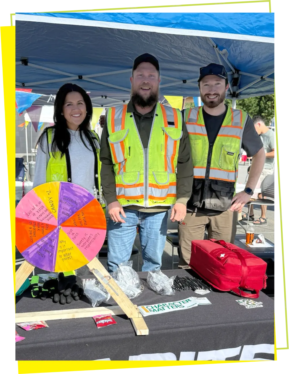 Three adults, two men and one woman, are standing behind a table at an outdoor event, possibly a safety fair. The two men and the woman are all smiling and wearing safety vests. A prize wheel, a red first-aid kit, and other promotional items are on the table in front of them. The man in the middle has a beard and is wearing a green shirt. A person is visible in the background, and there is a blue tent overhead.