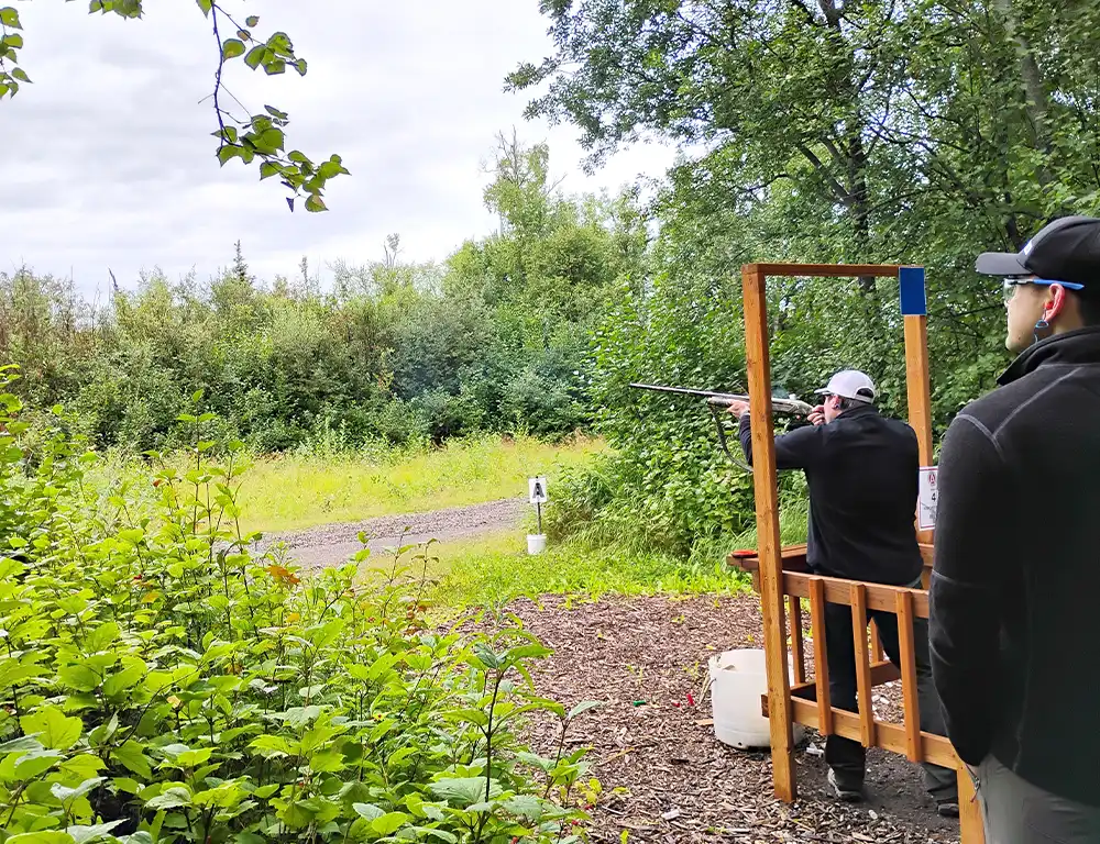 Side view of two men on a wooden platform at a sporting clays course, one man aiming a shotgun