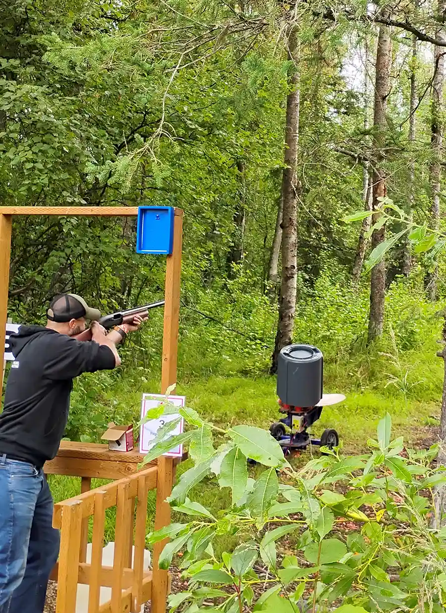 Man in black hoodie and jeans shooting a shotgun at a clay target in a wooded outdoor range.