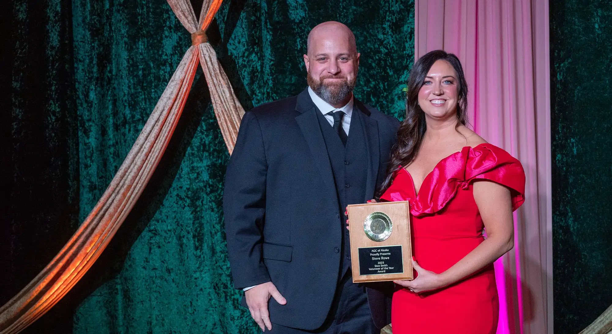 Award presentation at an AGC of Alaska gala featuring a man in a black suit and a woman in a red gown holding a wood and gold recognition plaque.