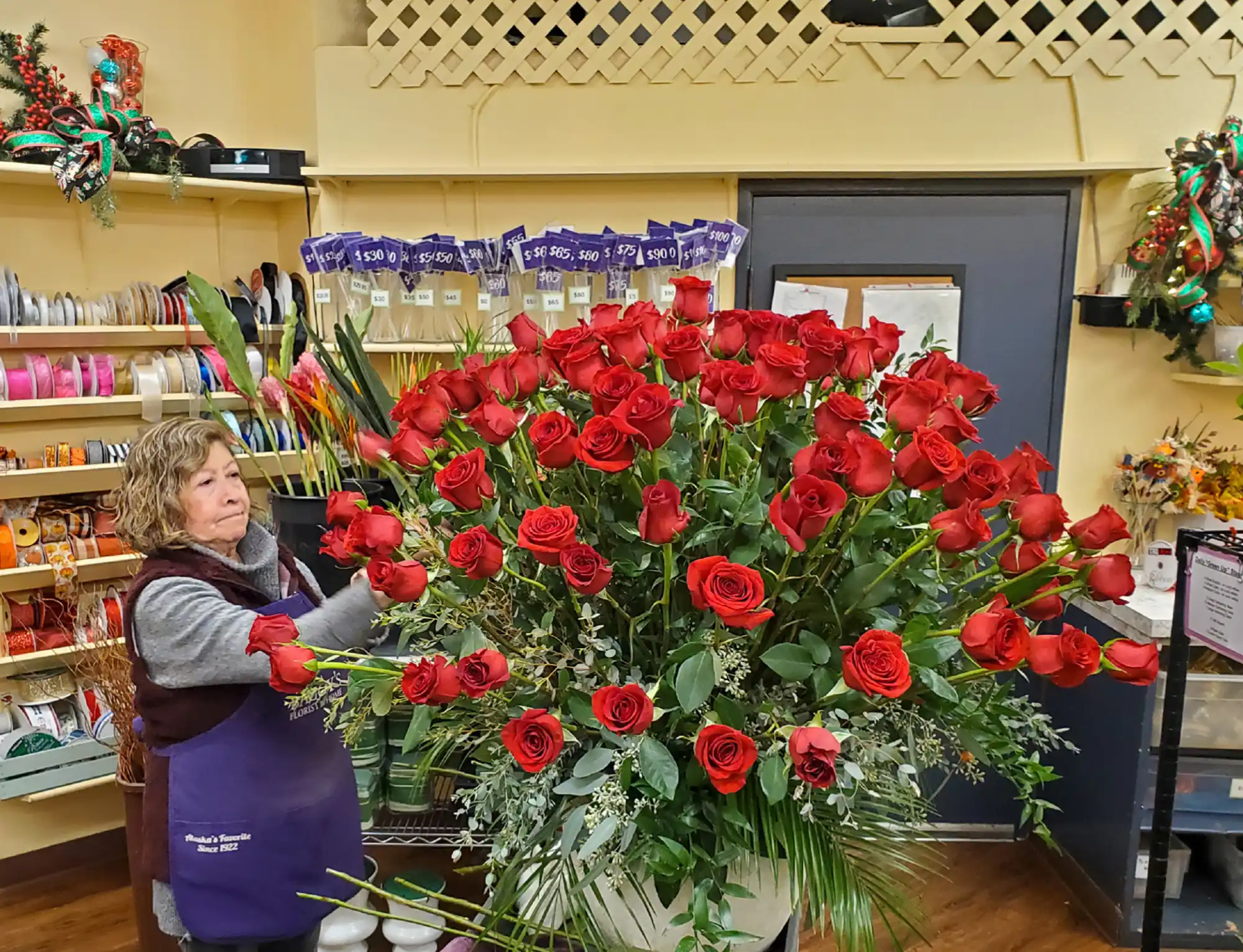 A florist in a purple apron arranges a massive, dense bouquet of dozens of long-stemmed red roses in a flower shop.