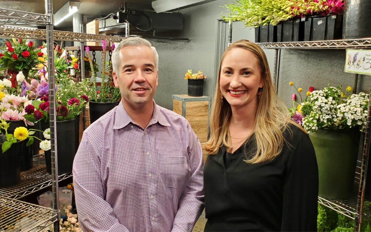 Adam Baxter and Kristen Keifer standing together in a flower shop surrounded by floral arrangements.