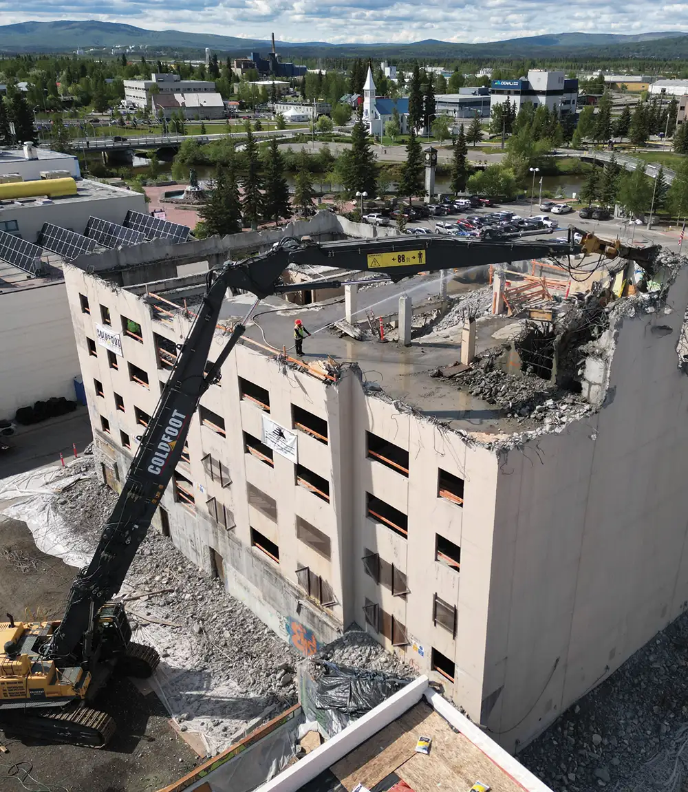 A high-reach demolition excavator with a long black arm tears into the top floor of a multi-story concrete building while a worker sprays water to control dust on the roof.