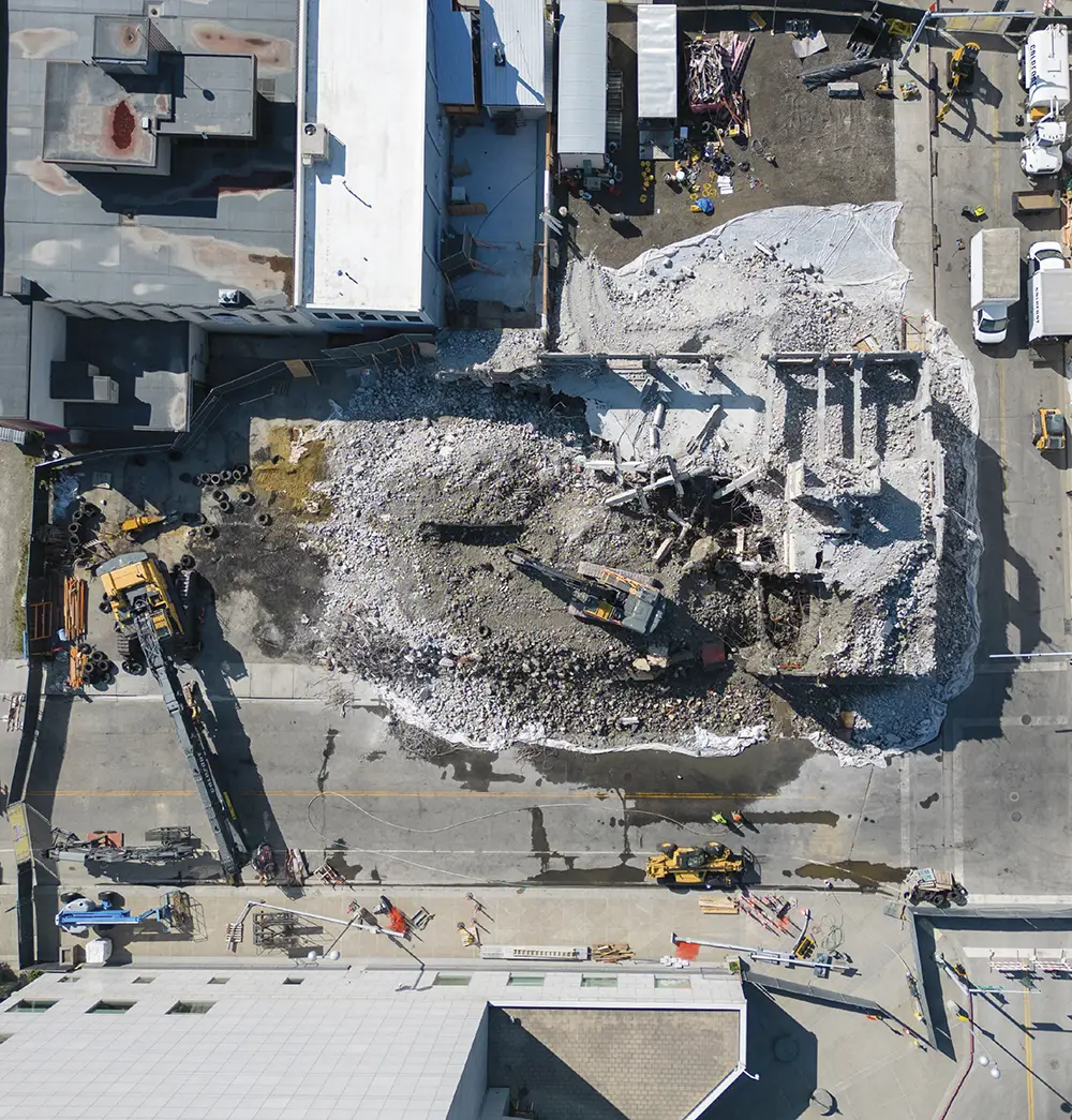 Direct overhead aerial view of a building demolition site showing an excavator surrounded by concrete rubble, debris piles, and white protective tarps spread across the ground.