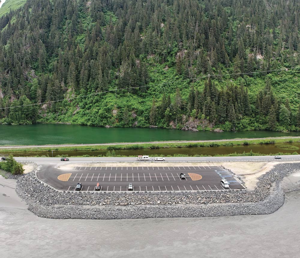 High-angle view of a newly paved and striped parking lot built on a rock-reinforced embankment next to a highway and river, with a lush green forest on the opposite bank.