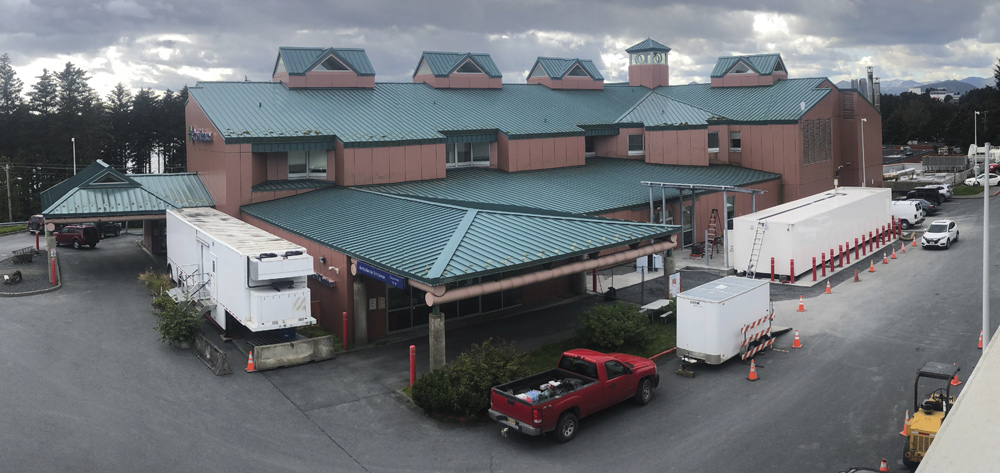 A wide shot of a salmon-colored medical building with a green metal roof, featuring several white mobile medical units and construction trailers parked in the adjacent lot.
