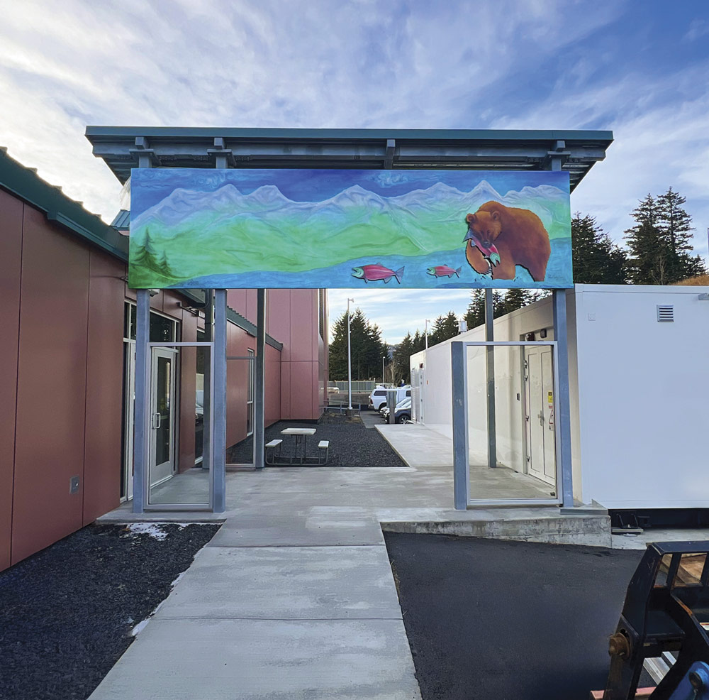 A vibrant outdoor mural depicting a brown bear catching a salmon in a mountain river is displayed above a concrete walkway between two sections of a medical facility.
