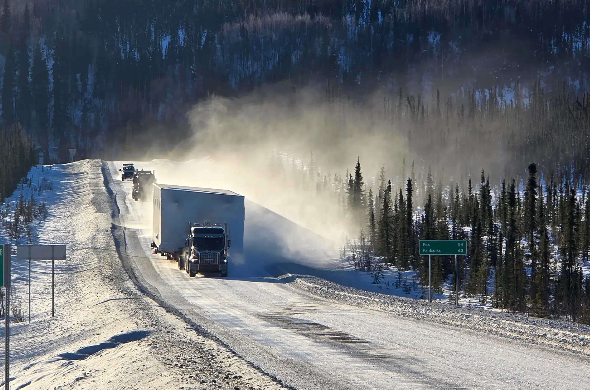 A large transport truck hauling a massive, rectangular white module on a snow-covered highway flanked by evergreen trees, with a cloud of white dust rising behind it.