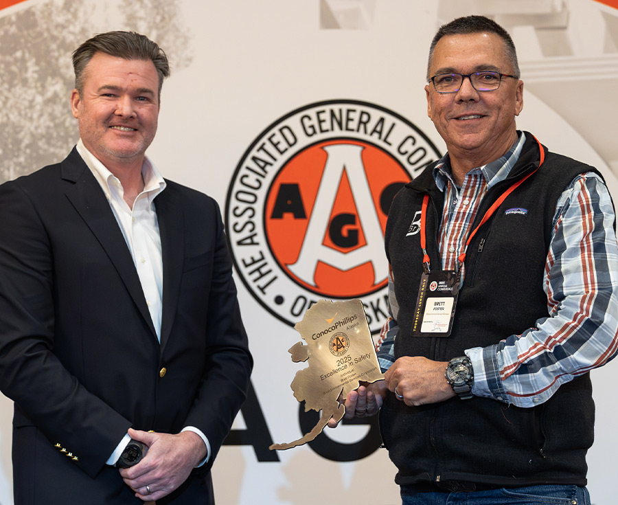 Jason Charton and Brett Foster posing with a gold, Alaska-shaped safety award in front of an Associated General Contractors (AGC) of Alaska backdrop.