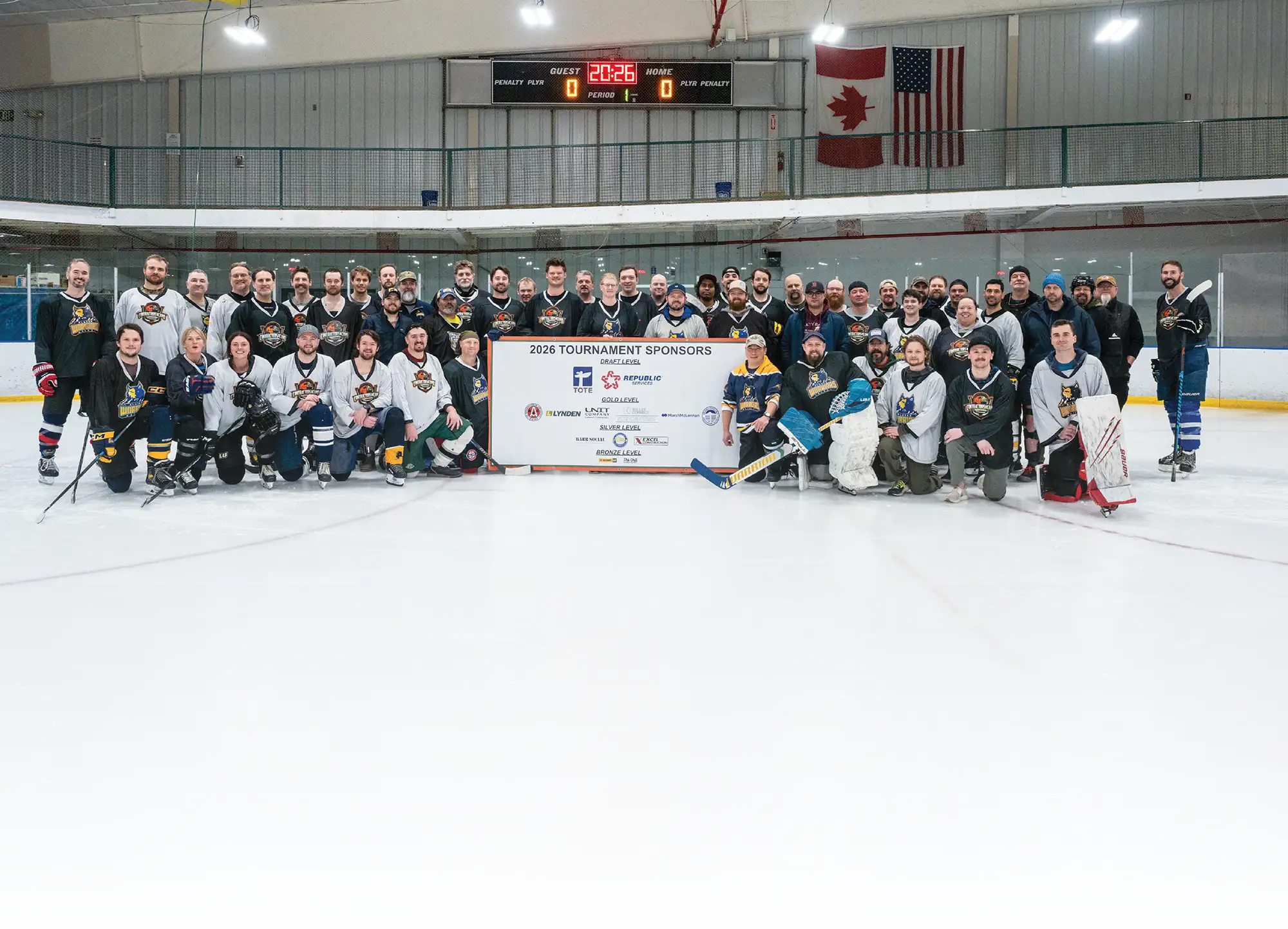 Large group of hockey players in black and white jerseys posing behind a "2026 Tournament Sponsors" sign on an ice rink with American and Canadian flags.