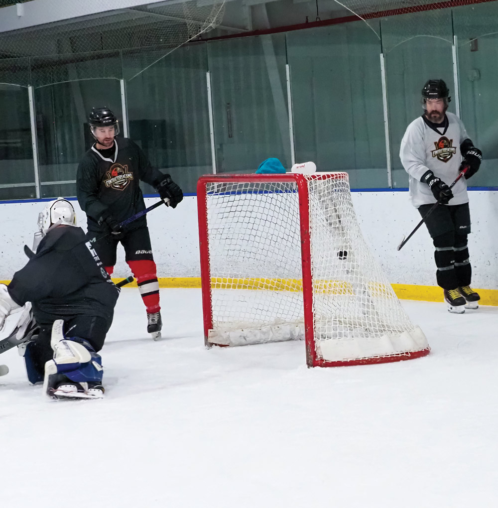 A hockey goalie in blue and white pads makes a save while opposing players in black and white jerseys look for a rebound near the red goal frame.