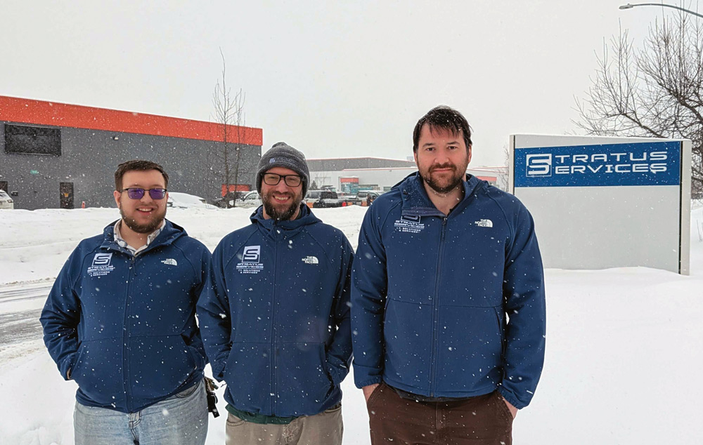 Three men in matching blue Stratus Services jackets stand in the snow in front of a company sign. The weather is lightly snowing, and a grey and orange building is in the background.