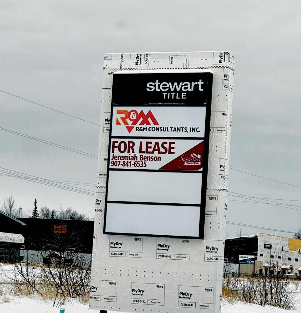 A commercial pylon sign wrapped in white protective housewrap features logos for Stewart Title and R&M Consultants, along with a "For Lease" contact sign in a snowy setting.