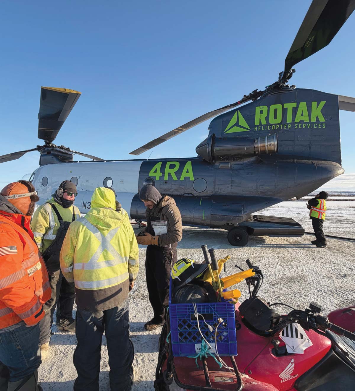 Workers in winter clothing stand before a large ROTAK Helicopter Services Chinook on a snowy landing pad.