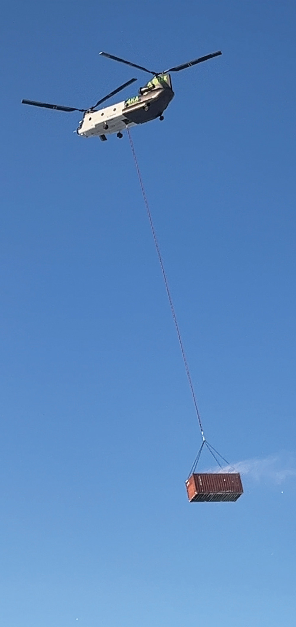 A large Chinook helicopter transports a brown shipping container via a long cable against a clear blue sky.