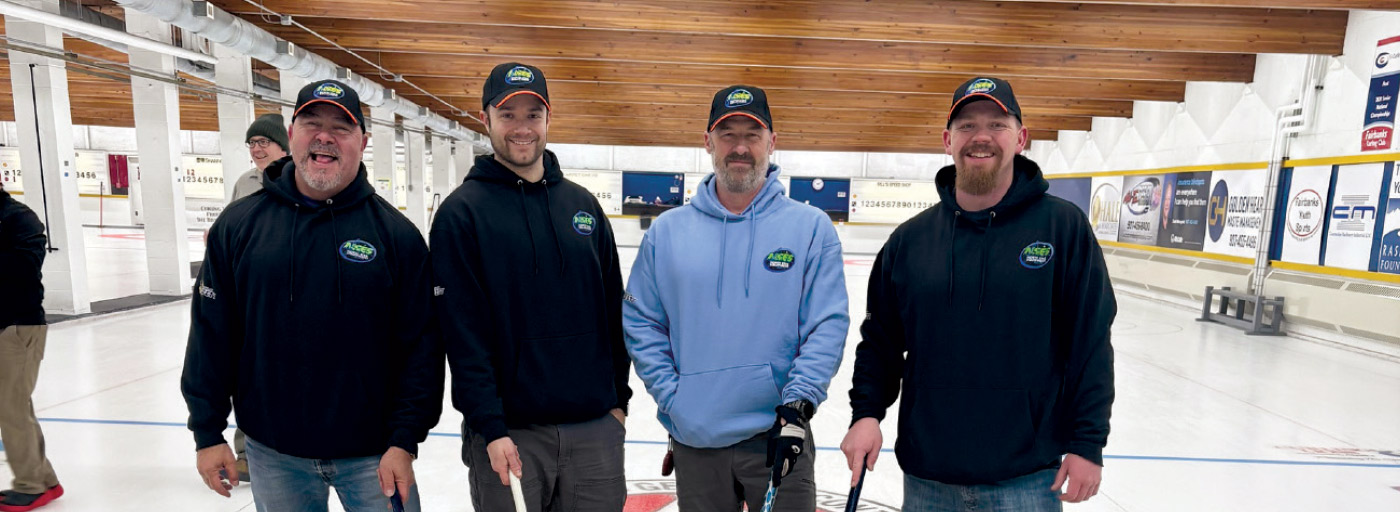 Four men in matching hoodies and hats pose on a curling ice rink during a community sporting event.