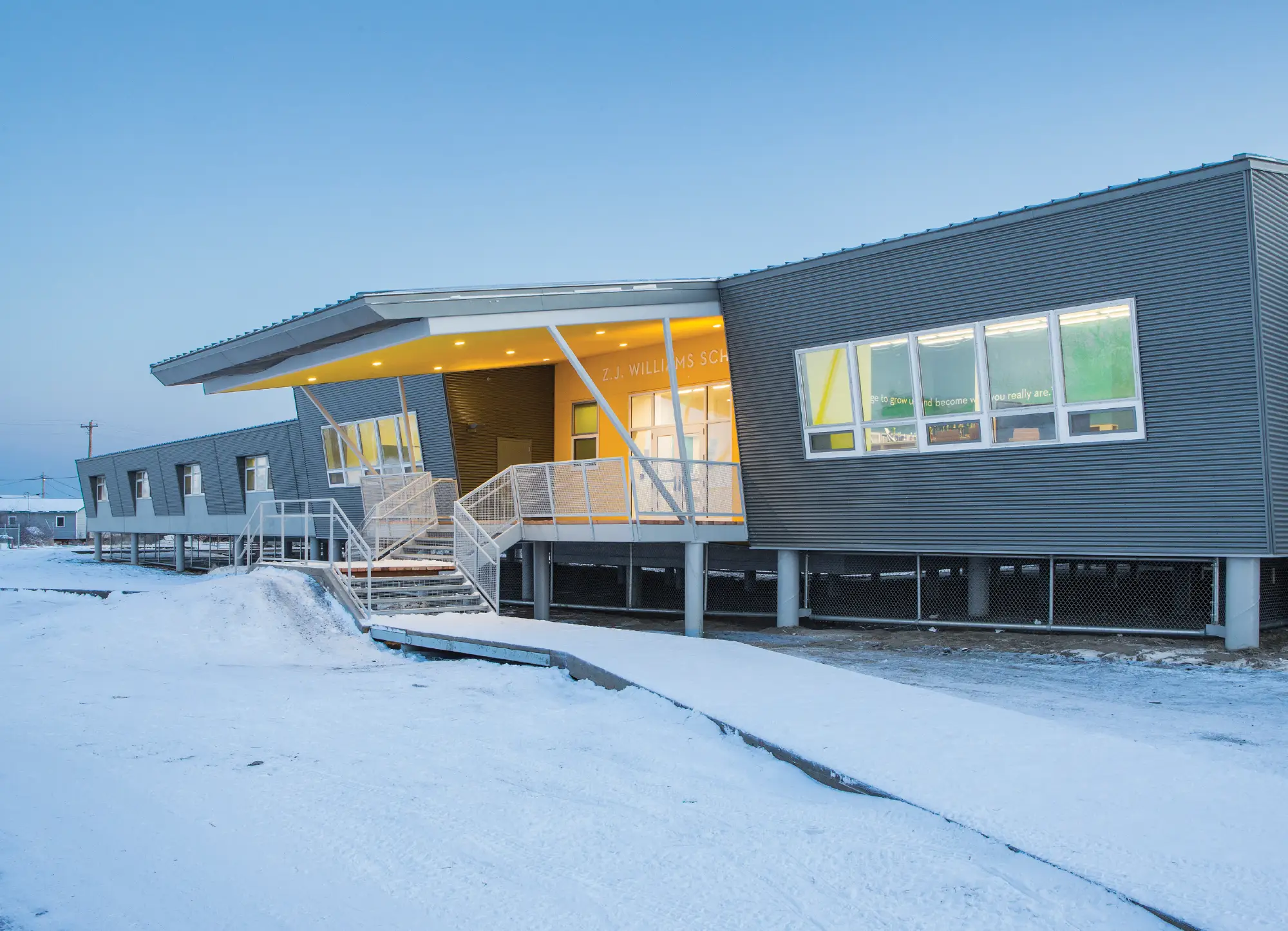 Modern grey and yellow Z.J. Williams School building built on stilts in a snowy Alaskan landscape under a clear blue sky.