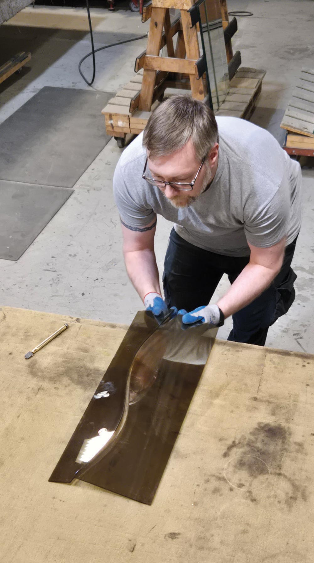 A man wearing blue gloves scores a long piece of tinted glass on a workbench in a professional workshop.