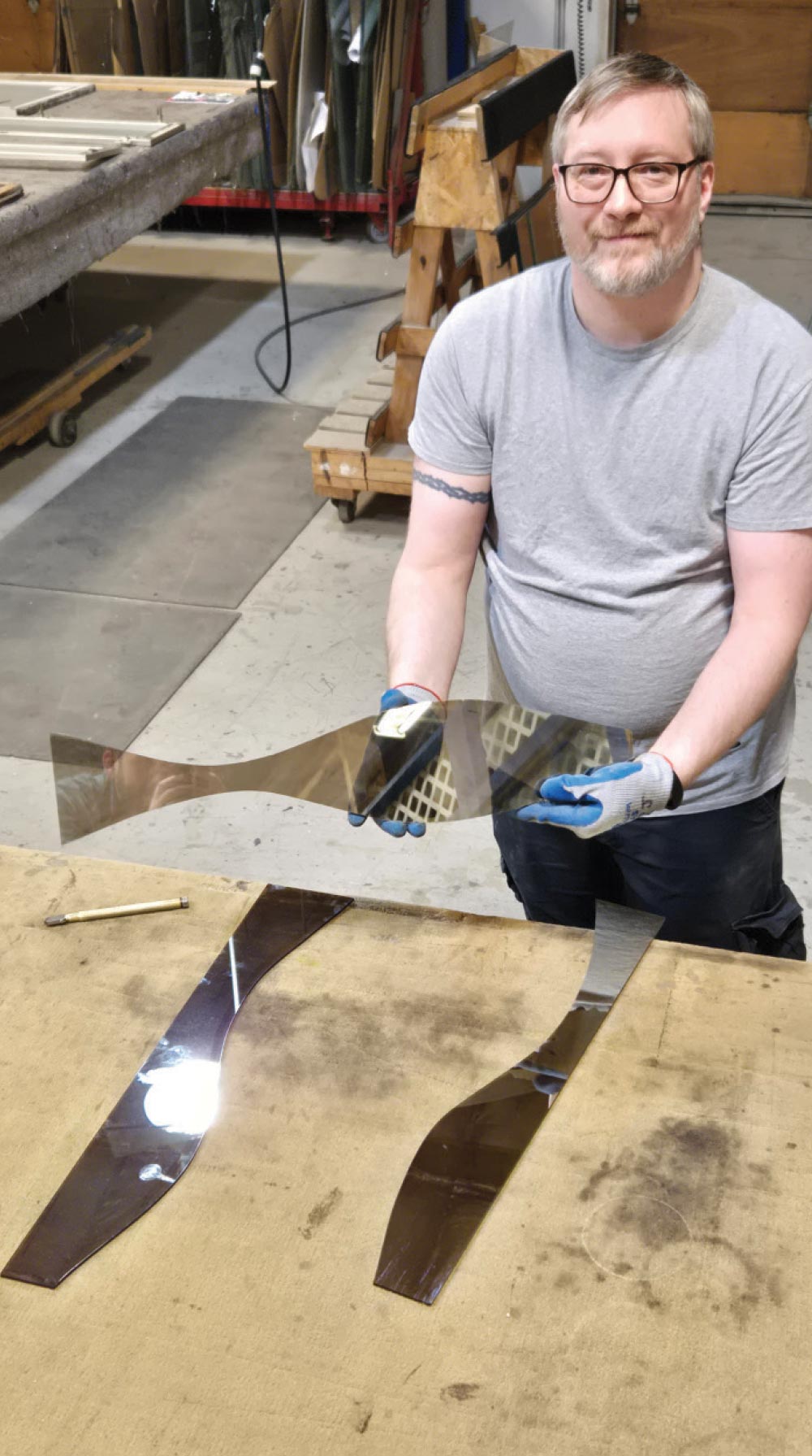 A man in a workshop holds up a custom-cut, curved piece of tinted glass, with other similar pieces on the table below.
