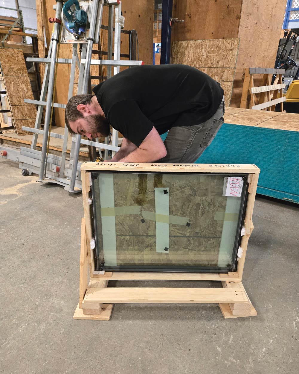 A worker in a black shirt assembles a window unit secured in a wooden frame labeled "Arctic Slope Native Association."