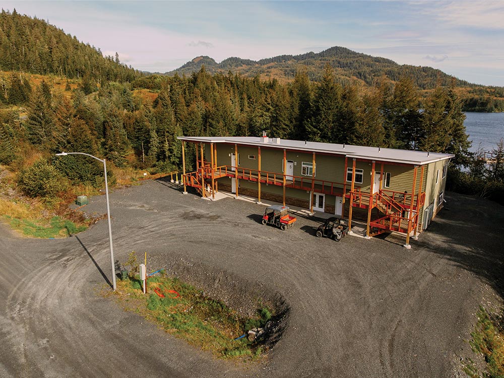 A two-story green lodging facility with wooden balconies and stairs sits on a gravel lot. Two utility vehicles are parked in front. Forested hills and a body of water are visible in the background.