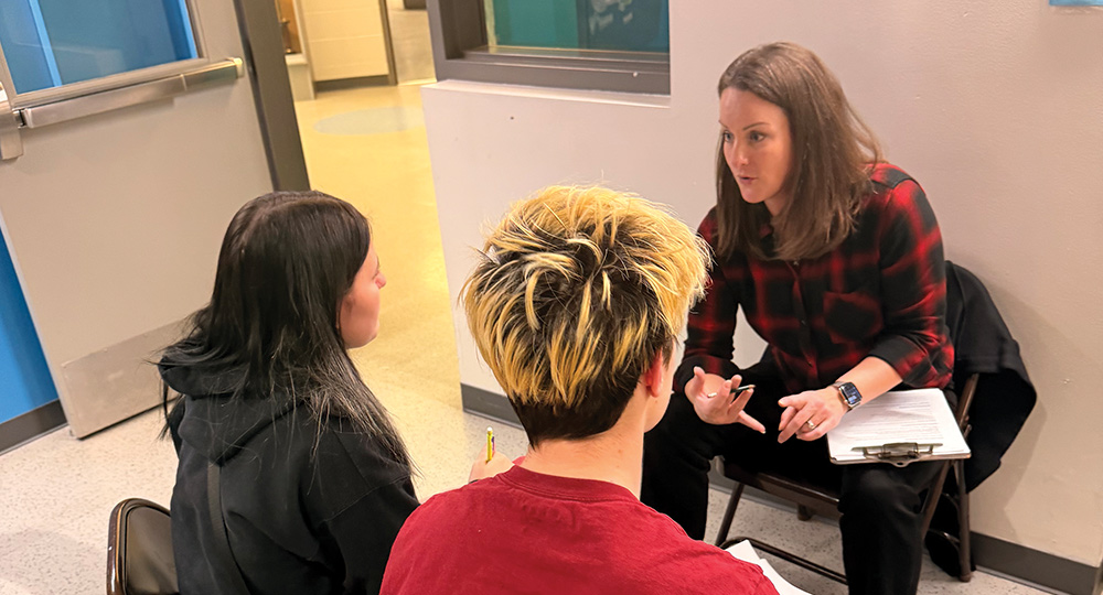 A woman in a red and black flannel shirt gestures and speaks to two young people, one with distinctive bleached hair, sitting in front of her. She holds a clipboard on her lap.